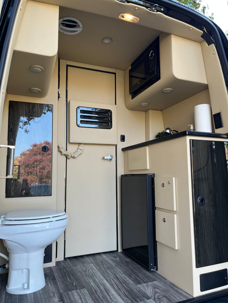Modern RV rental interior with compact kitchen (microwave, fridge, sink) and a toilet. Beige & wood tones for comfortable travel.
