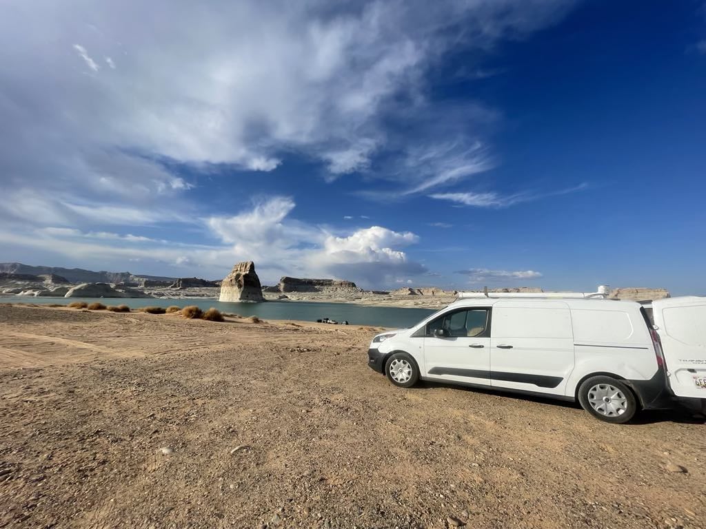 White RV rental van by a desert lake. A lone rock spire rises from the water, under a vast blue sky with clouds.