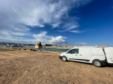 White RV rental van by a desert lake. A lone rock spire rises from the water, under a vast blue sky with clouds.
