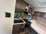 RV rental interior showcasing a compact kitchen area with a sink, stove, microwave, and an adjacent bed.