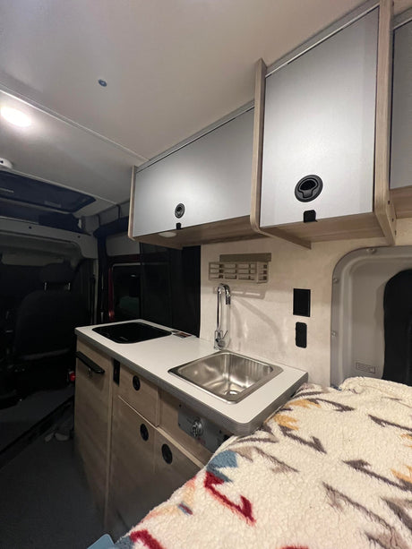 A well-equipped RV rental interior showing a kitchen with sink and stovetop, overhead storage, and a comfortable bed.