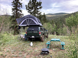 Dark green truck with roof-top tent deployed in a forest campsite. Chairs, table, and cooking gear ready. A compact outdoor RV rental alternative.