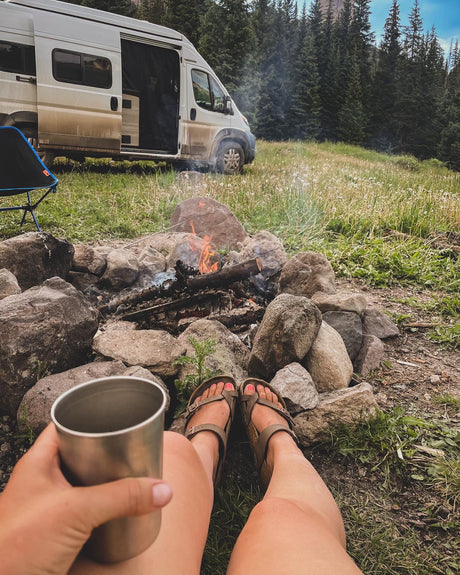 Enjoying a campfire from a ground-level view, feet propped up, with a dirty white camper van (RV rental) and trees in the background.