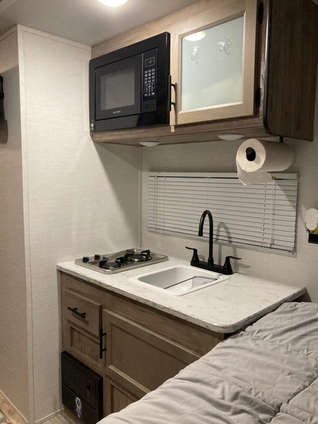 RV rental kitchen view: microwave, stove, sink, and counter space. Overhead cabinets, paper towel holder, and bed visible.