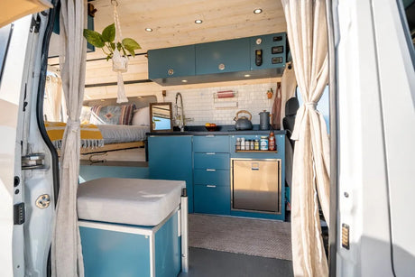 Explore this compact RV rental! A bright camper van interior features a blue kitchen with sink, fridge, and a cozy bed area.