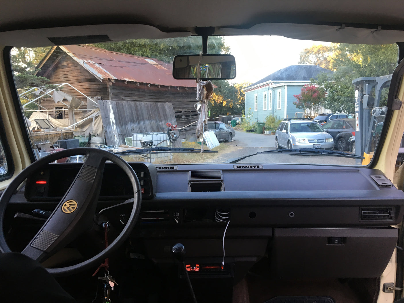 View from an RV rental dashboard, showing a steering wheel, rustic barn, blue house, and various vehicles parked outside.