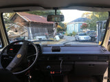 View from an RV rental dashboard, showing a steering wheel, rustic barn, blue house, and various vehicles parked outside.