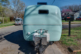 Rounded light blue camper rental with propane tanks on a residential street. A van and mailbox 1406 are visible.