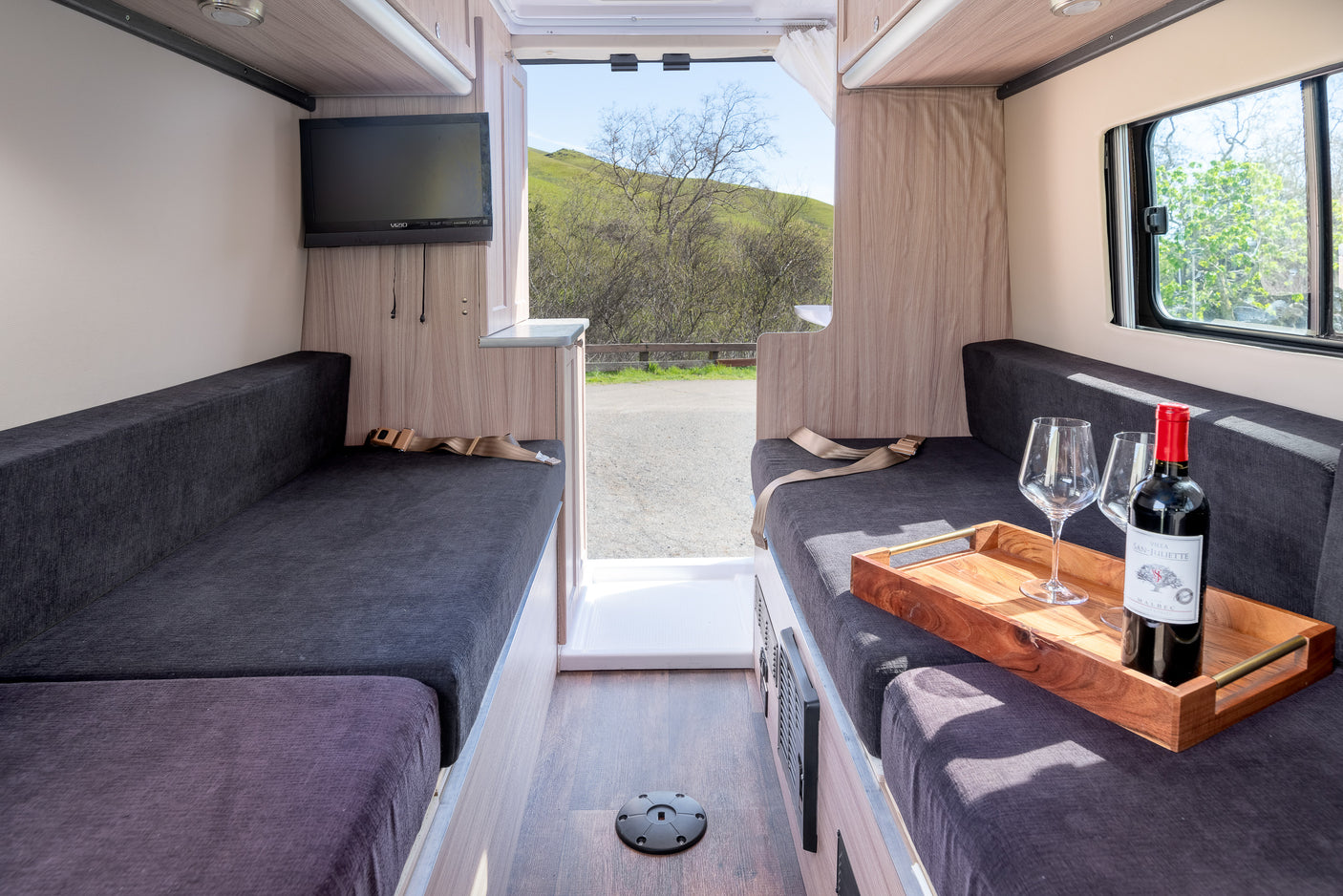 This rv rental offers a comfortable interior with dark upholstered seating, TV, wine setup, and scenic outdoor views.