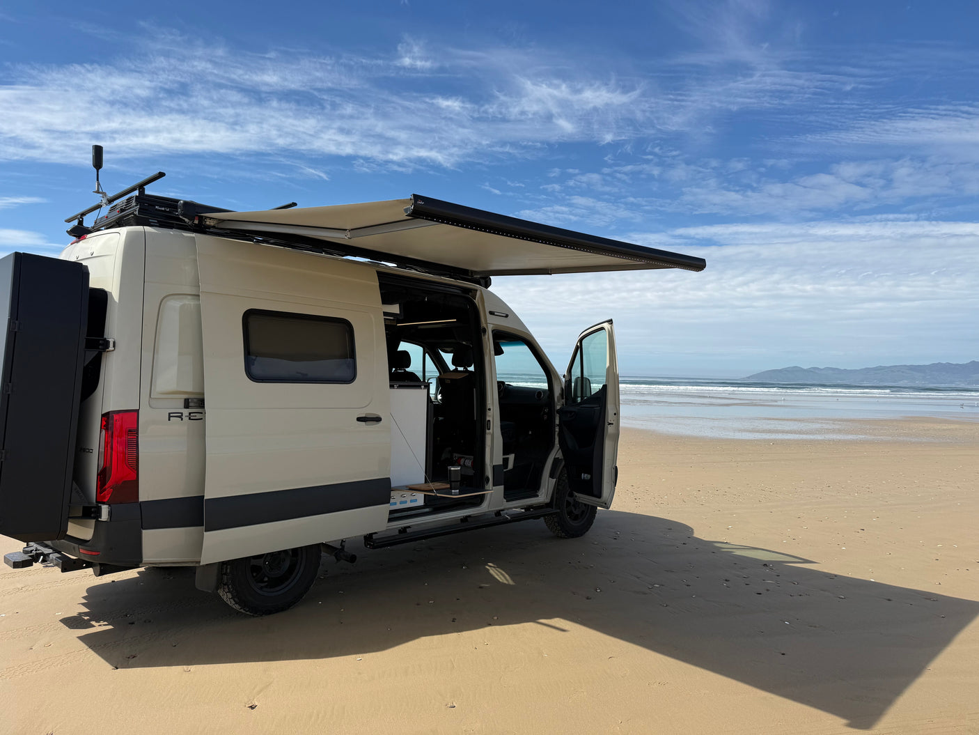 A cream rv rental camper van sits on a sandy beach with an extended awning, ocean, and mountains. Ready for adventure!