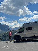 Excited traveler with arms up beside an RV rental, set against stunning mountains & blue sky. Perfect for outdoor adventure!
