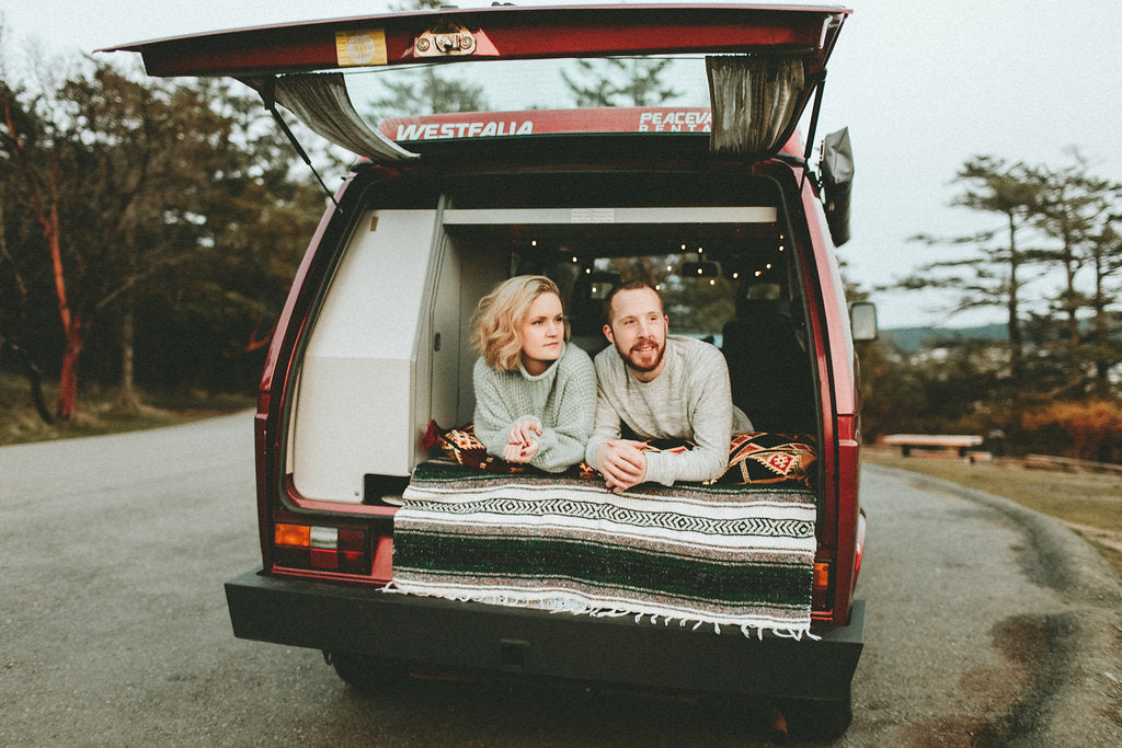 Two people relax on blankets in the open tailgate of a red Westfalia camper van. Perfect for an RV rental adventure or road trip.