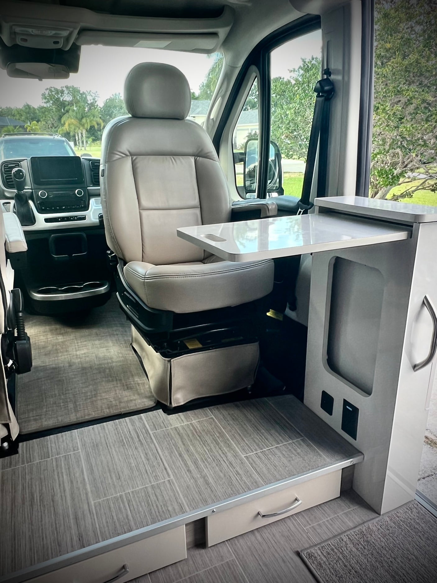 This RV rental interior features a beige swiveling driver's seat, a pull-out white table, and sleek wood-look tiled flooring.