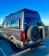 This custom dark gray camper van, an ideal RV rental option, features a roof rack, ladder, and rear-mounted spare tire for adventure.