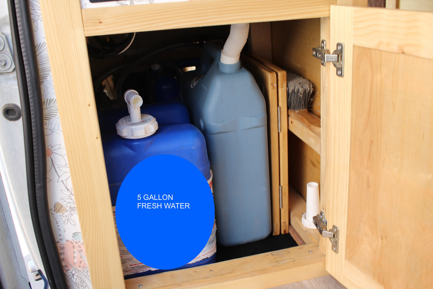 RV rental: Inside a wooden cabinet, a 5-gallon blue fresh water tank and a lighter blue water jug are stored, essential for travel.