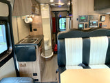 Inside an RV rental: view of the modern kitchen with stove/sink, dinette seating, and compact bathroom, plus control panels.