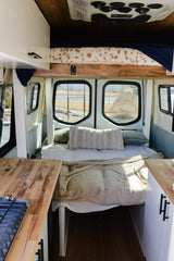 Inside a modern RV rental: a comfy bed with pillows, large windows overlooking the outdoors, and wooden kitchen counters.