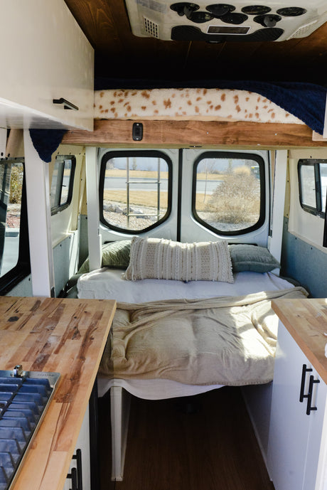 Inside a modern RV rental: a comfy bed with pillows, large windows overlooking the outdoors, and wooden kitchen counters.