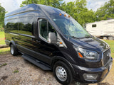 Stylish black Ford Transit van, suitable for rv rental adventures, parked near a white RV amidst trees.