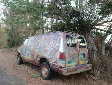 Psychedelic custom-painted van, like a unique RV rental, parked off-road by trees.