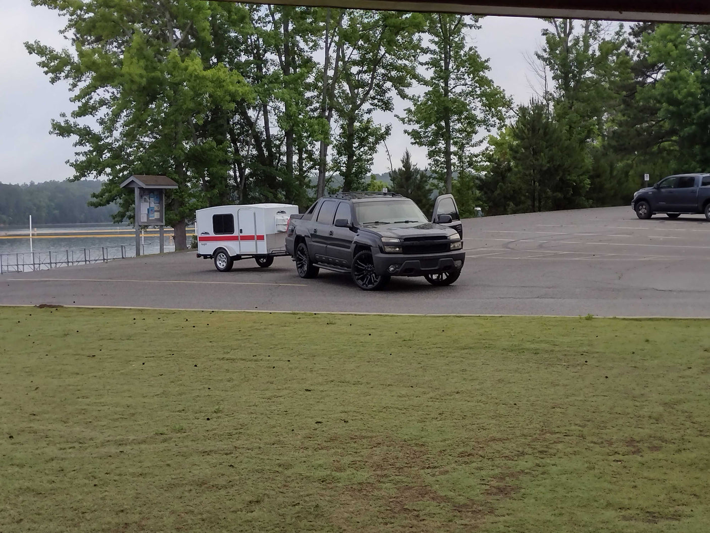 Dark truck towing a white rv rental camper trailer in a paved lot by a lake, surrounded by trees.