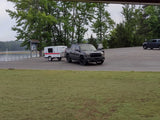 Dark truck towing a white rv rental camper trailer in a paved lot by a lake, surrounded by trees.