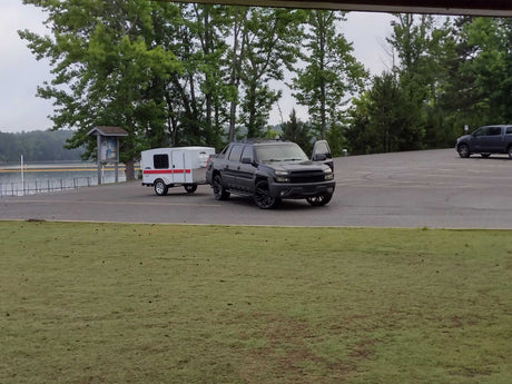 Dark truck towing a white rv rental camper trailer in a paved lot by a lake, surrounded by trees.
