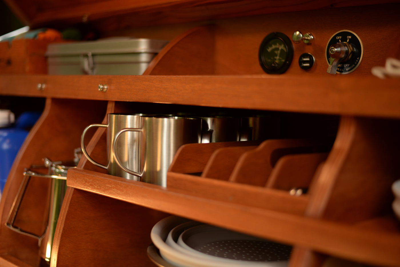 Compact RV rental interior showing organized wooden storage. Features battery controls, stainless mugs, & kitchenware for mobile living. — photo 6