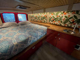 RV rental interior: bed, vibrant tropical curtains, red cabinets, sink. Woven ceiling & blurred windows add charm.