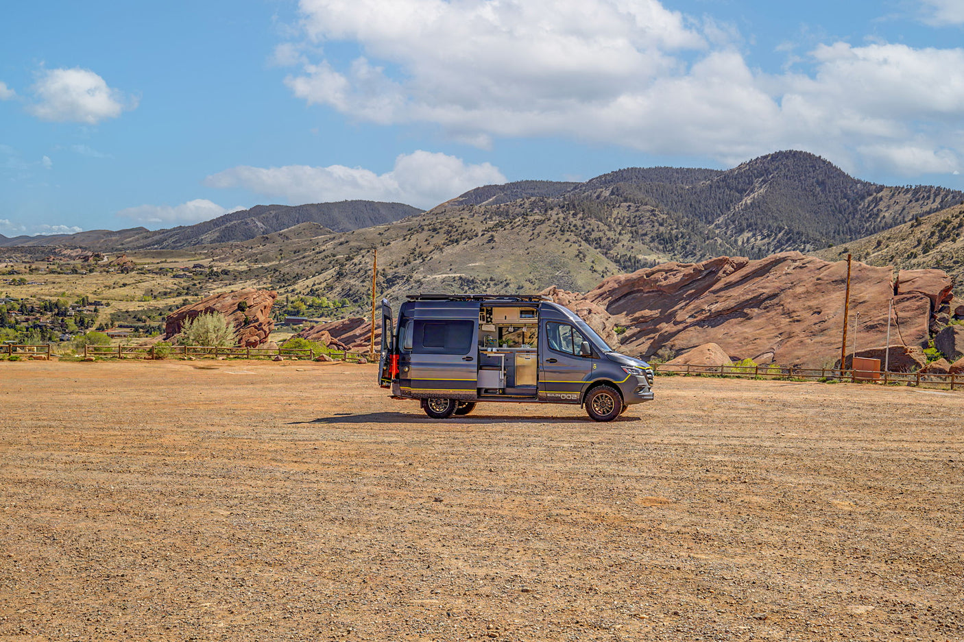 Modern grey camper van, an RV rental, open to its interior, parked on a gravel lot with scenic red rocks and mountains beyond.