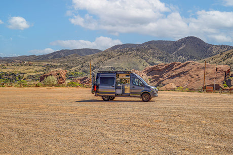 Modern grey camper van, an RV rental, open to its interior, parked on a gravel lot with scenic red rocks and mountains beyond.