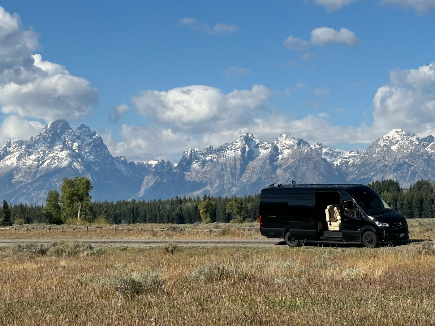Black RV rental van parked by a road, revealing its interior, with majestic snow-capped mountains & forests in the background.