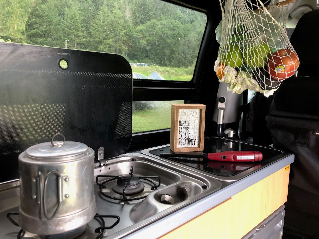 Cozy RV rental kitchenette with "Tacos" sign, fresh fruit, and rainy forest views. Perfect for camping.