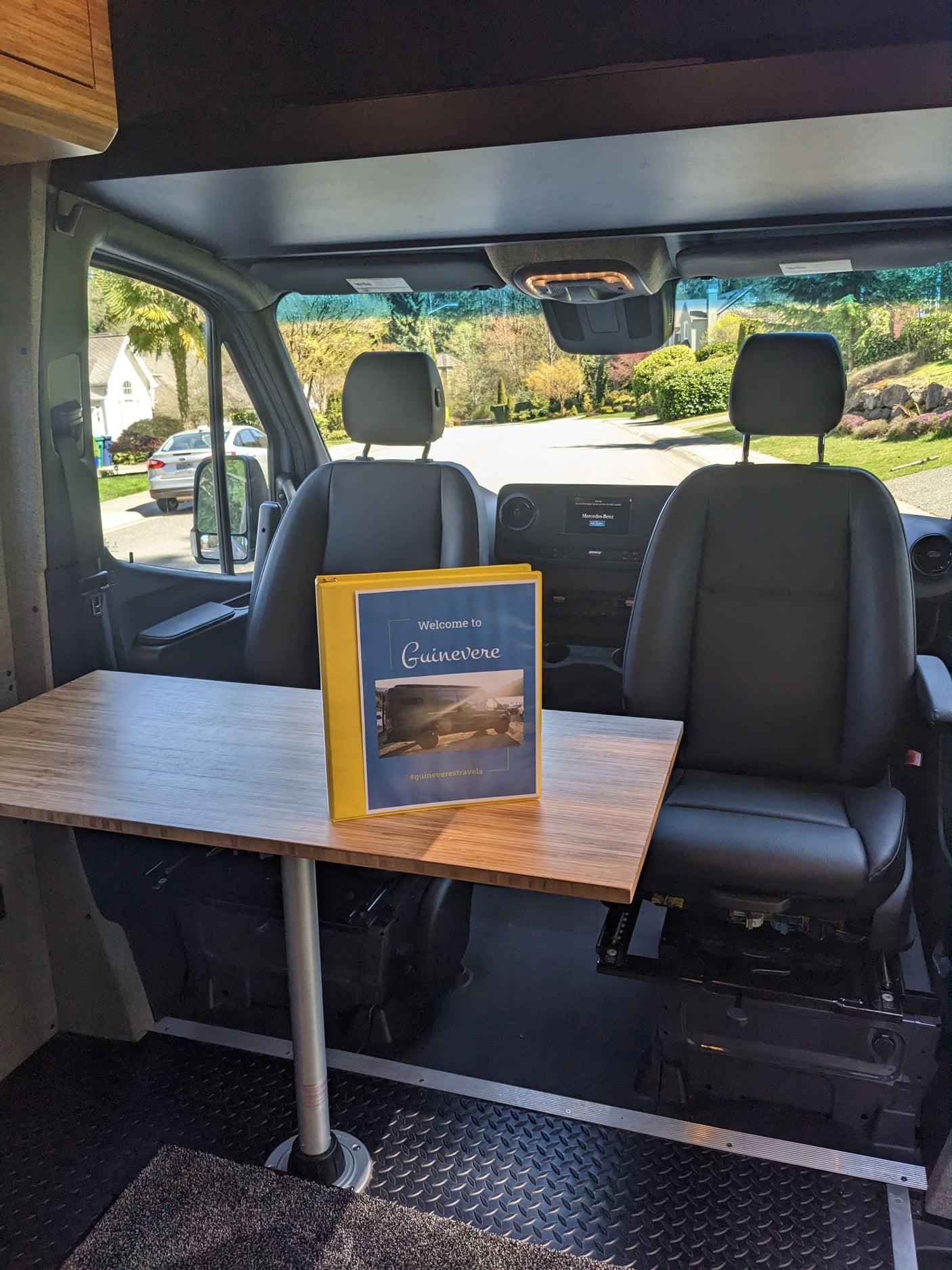 Inside a spacious RV rental: front seats, a wooden table with a welcome binder, and a view of a quiet residential street.