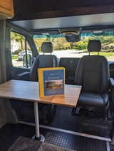 Inside a spacious RV rental: front seats, a wooden table with a welcome binder, and a view of a quiet residential street.