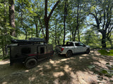 Electric truck tows an off-road camping trailer through a sunny forest. Great for an RV rental adventure!