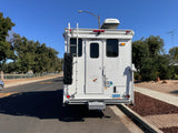 White truck camper, great for an RV rental, parked on a sunny street. Features a ladder, recovery boards, Yakima roof box, Nevada plate.