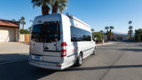 A silver Airstream Interstate RV rental van, with a bike rack, parked on a sunny desert street lined with palm trees.