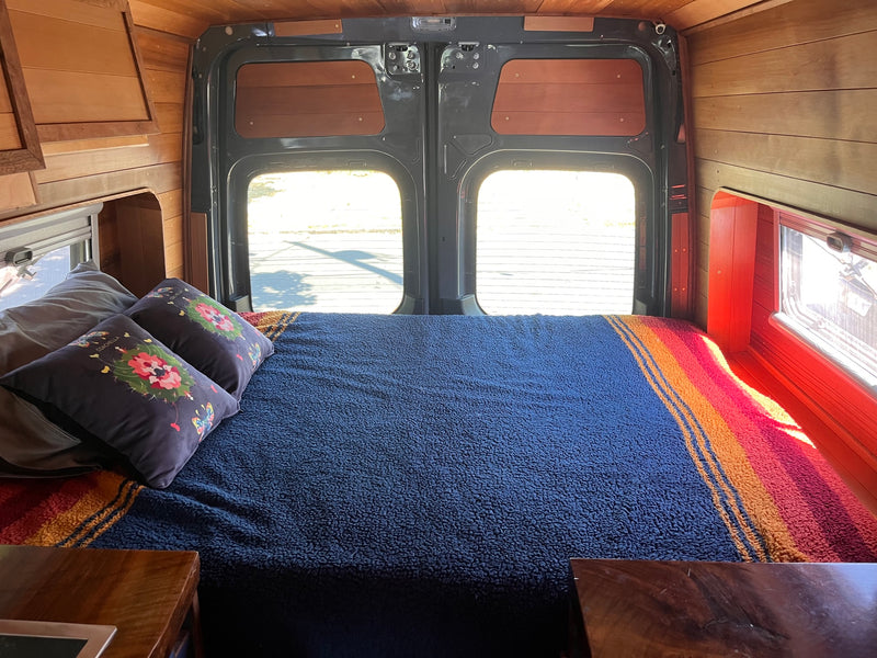 Cozy RV rental interior: warm wood walls, a large bed with colorful blankets, and a bright view out the back. — photo 5