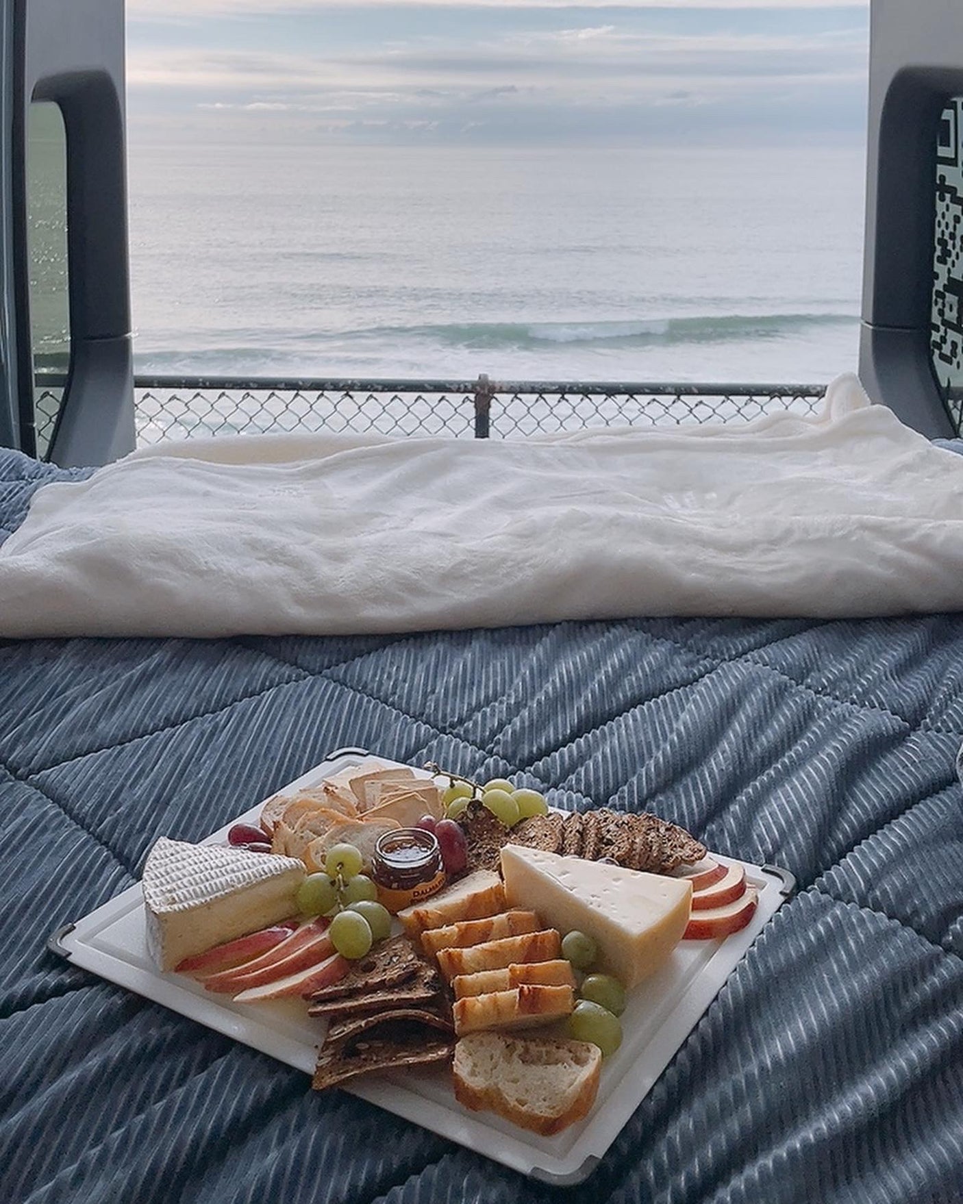 RV rental comfort: A cozy bed and charcuterie board with ocean views from an open van. Perfect travel escape!