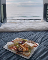 RV rental comfort: A cozy bed and charcuterie board with ocean views from an open van. Perfect travel escape!
