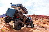 Black off-road trailer with rooftop tent, ideal for an RV rental adventure, navigates red rocky terrain behind a Jeep.