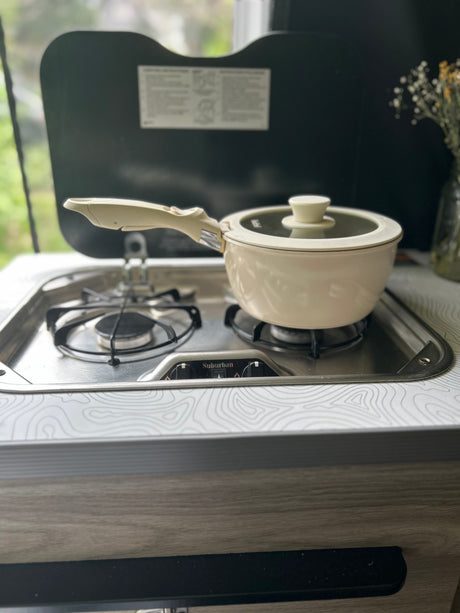 Cooking a meal in an RV rental, featuring a compact Suburban cooktop with a cream pot. Ideal for travel adventures.