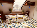 Cozy RV rental interior: patterned bed with vehicle theme, accent pillow, and window view of a USPS truck.