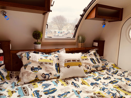 Cozy RV rental interior: patterned bed with vehicle theme, accent pillow, and window view of a USPS truck.