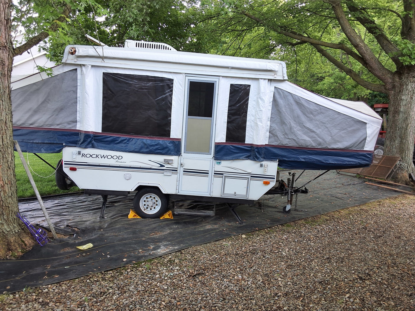 A white Rockwood pop-up camper, perfect as an RV rental or travel trailer, is set up on a black tarp among trees.