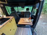 Inside a comfy RV rental: a built-in kitchenette, dining table with baguettes & whiskey, overlooking a serene forest path.