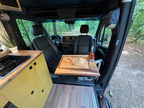 Inside a comfy RV rental: a built-in kitchenette, dining table with baguettes & whiskey, overlooking a serene forest path.