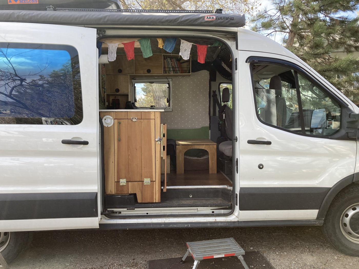 This white camper van, suitable for an rv rental, features an open side door revealing a custom wood interior & colorful prayer flags. — photo 8