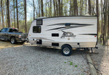 RV rental setup: A Jayco travel trailer hitched to a pickup truck on a gravel campsite amidst trees, perfect for camping.
